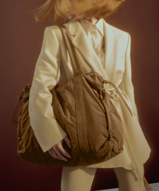 A person in a cream-colored blazer and matching trousers carries a large brown drawstring bag over their shoulder against a dark reddish-brown background.